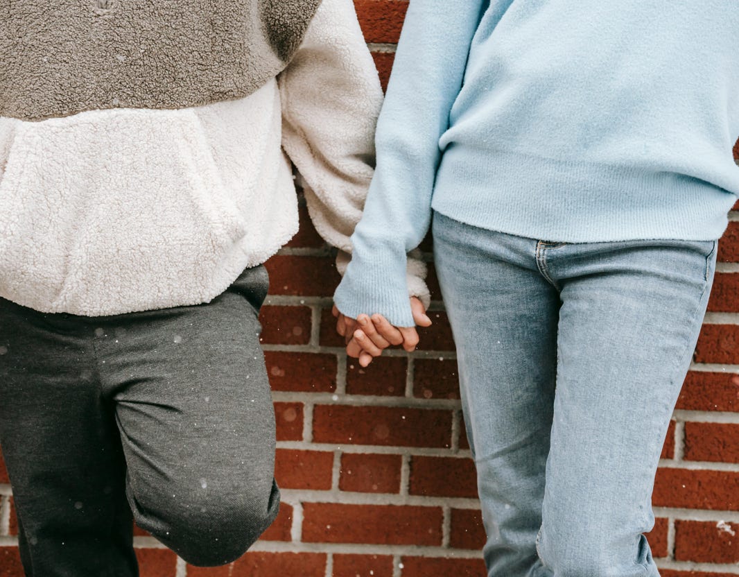 Crop unrecognizable couple in warm sweaters standing near brick wall of building and holding hands in snowy day