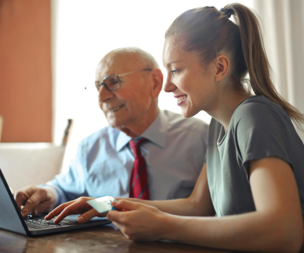 Young woman in casual clothes helping senior man in formal shirt with paying credit card in Internet using laptop while sitting at table